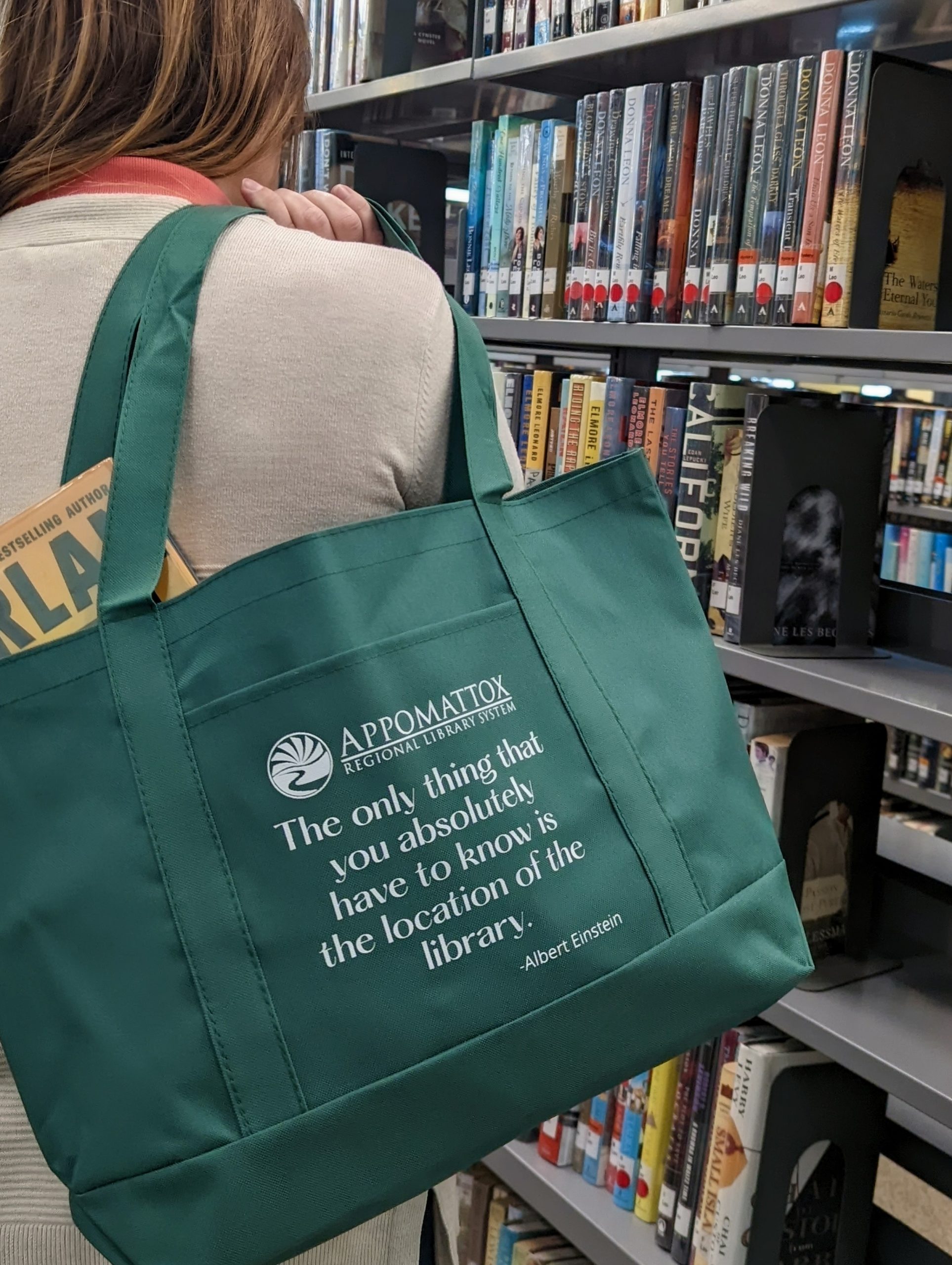 A woman standing in front of a library bookshelf carrying a green tote bag with a library quote on it. Peaking out of the bag is a book.